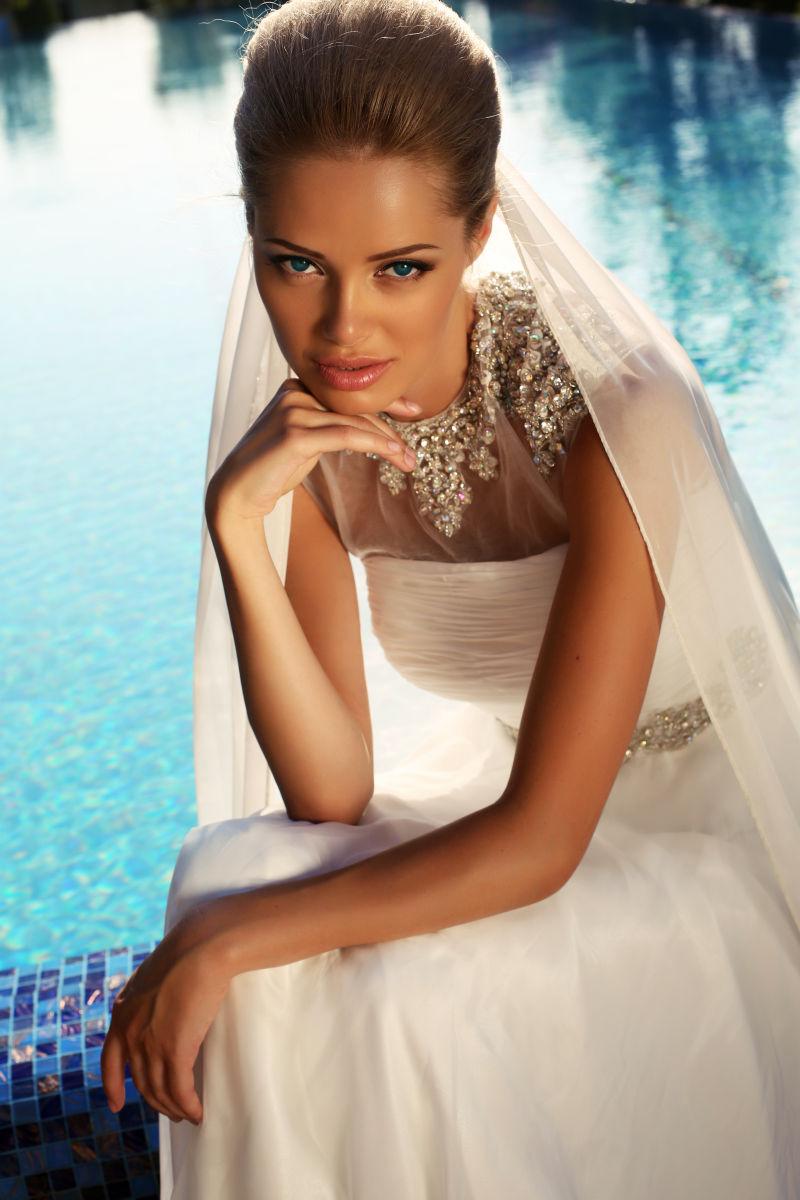 美丽的金发新娘穿着优雅的婚纱摆在游泳池旁