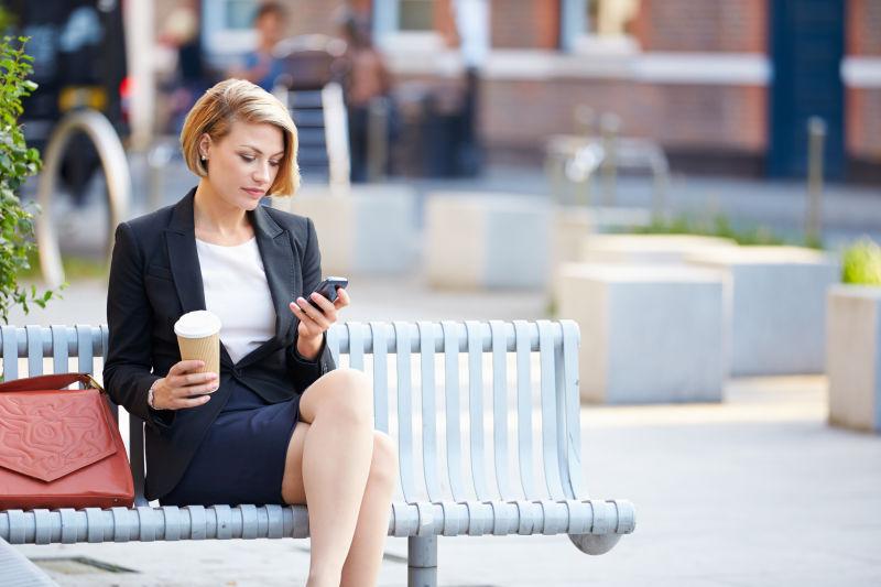 坐在公园长椅上的女商人用手机喝咖啡