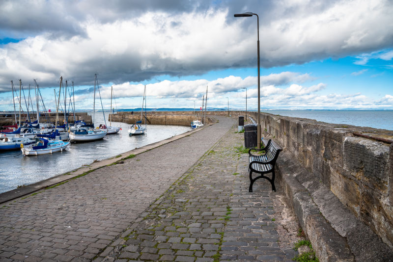 码头上一条鹅卵石铺就的港湾小道旁的空长椅照片的左边是帆船