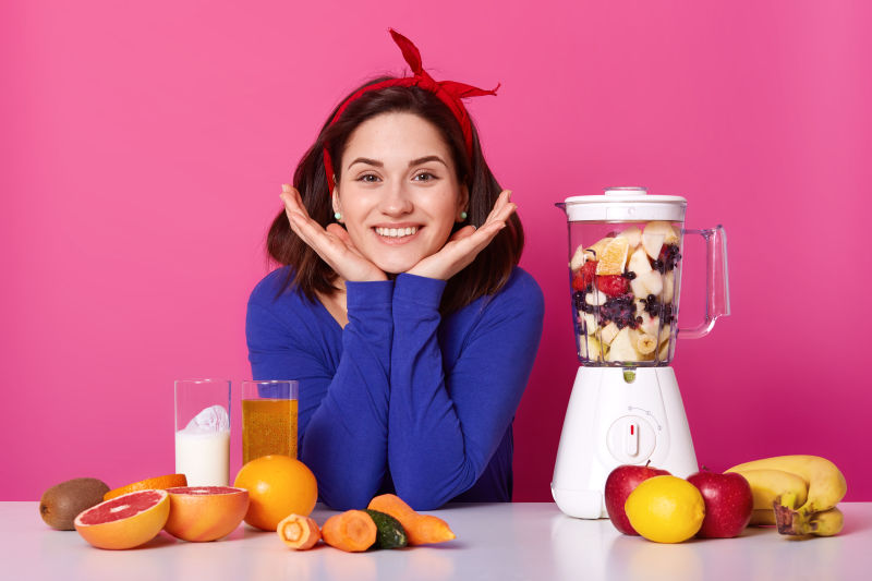 迷人的女孩手下巴戴红色头巾蓝色跳线使用食品加工机为新鲜不同的水果和蔬菜制作新鲜的冰沙节食健康饮食理念