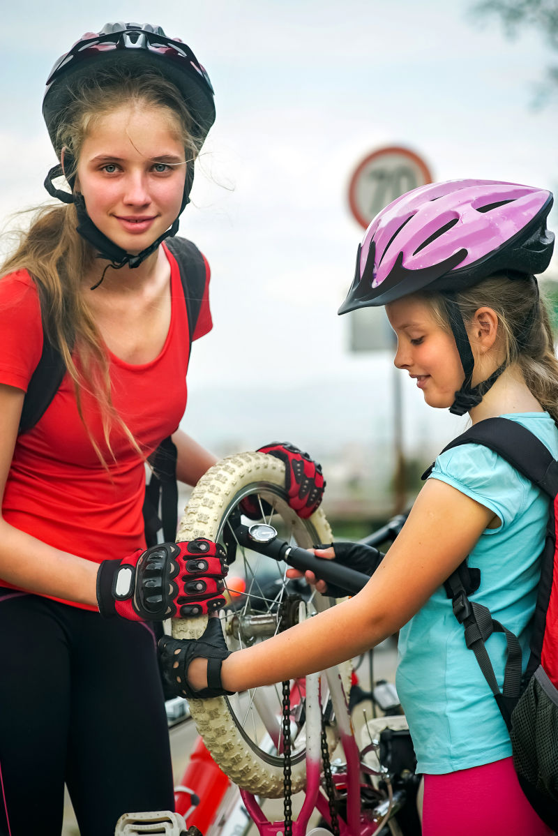 女孩们戴着自行车头盔和自行车用手摇泵女孩给自行车轮胎打气背景和天空的道路和交通标志
