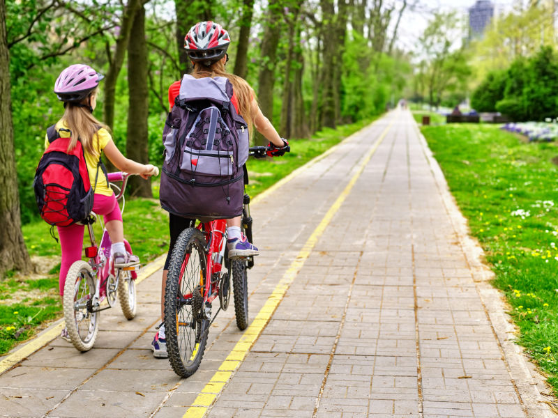 骑自行车的女孩女孩们戴着自行车头盔背着帆布包骑着自行车女童在黄色自行车道上骑车自行车共享计划节省金钱和时间后视图