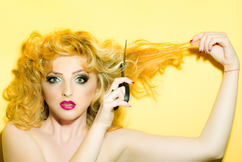 一位美丽性感的年轻复古金发女性的肖像卷发和鲜红的嘴唇向前看拿着剪刀背景是黄色水平的图片
