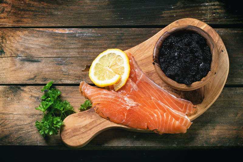一碗黑鱼子酱和几块腌鲑鱼放在橄榄木板上旧木桌上放着一把老式的刀子顶视图