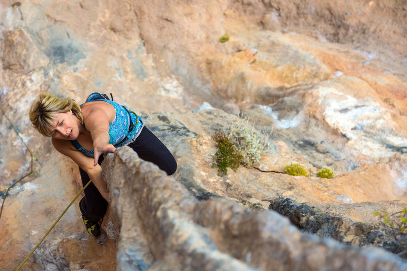 在岩石地形上的金发可爱的攀岩者使女性运动员很难移动-她们攀岩时自然的岩石保持住并悬挂在深深的深渊上