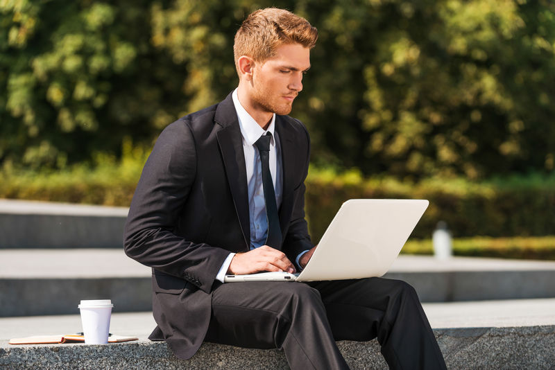 在户外工作-穿着衬衫和领带的自信的年轻人坐在户外用笔记本电脑工作