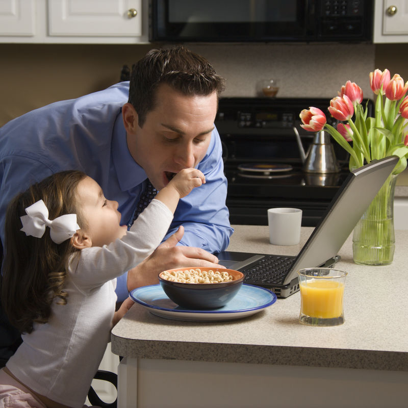 穿着西装的白人父亲用笔记本电脑和女儿在厨房给他吃早餐