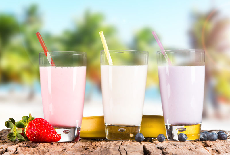 新鲜牛奶草莓蓝莓和香蕉饮料放在木桌上-配以新鲜水果和热带海滩背景的各种蛋白质鸡尾酒
