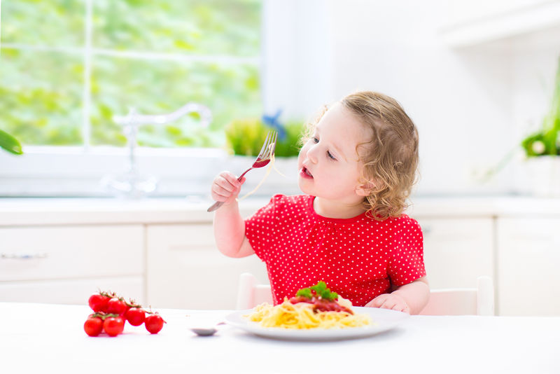 可爱的卷发的笑着的穿着红色衬衫的小女孩坐在一个阳光明媚的有大窗户的现代厨房里-用叉子和勺子玩着吃意大利面番茄酱和蔬菜来做健康的午餐