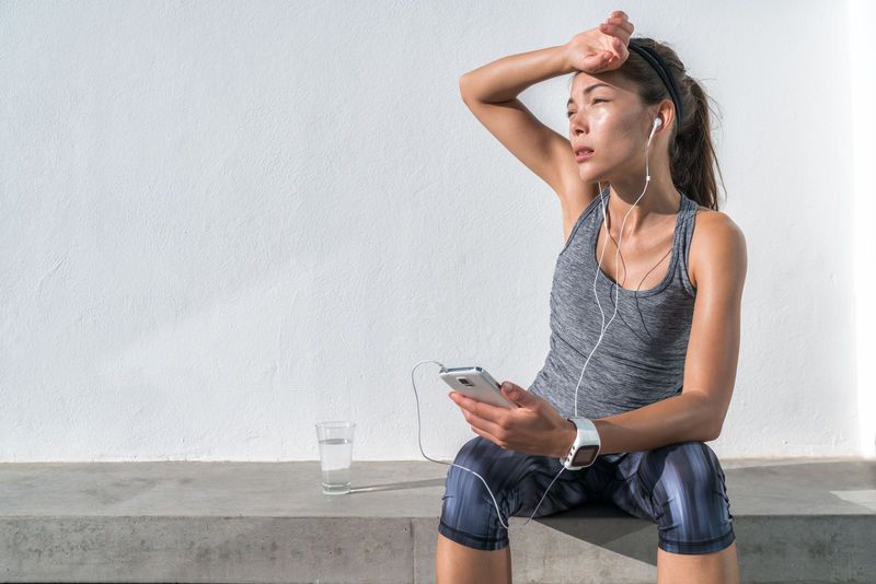 疲累的健身女性在训练困难后在电话上休息听音乐。精疲力竭的亚裔跑步者由于在健身房锻炼而脱水，感到精疲力竭和脱水。