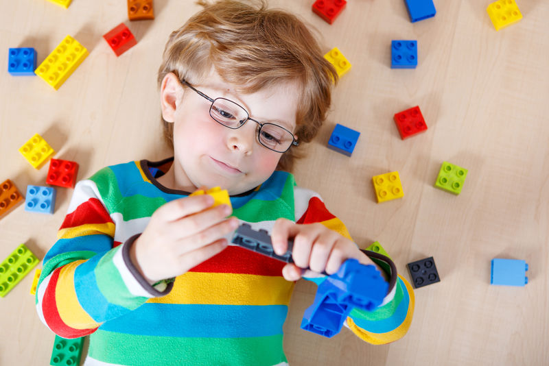 小金发男孩在室内玩着许多彩色塑料块-孩子穿着彩色衬衫和眼镜-玩建筑和创造