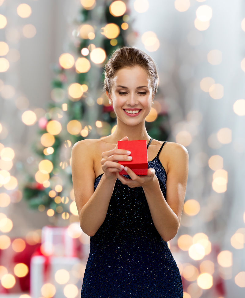 节日、庆祝活动和人们的观念-微笑的女人穿着衣服-在圣诞树灯的背景上拿着红色的礼品盒