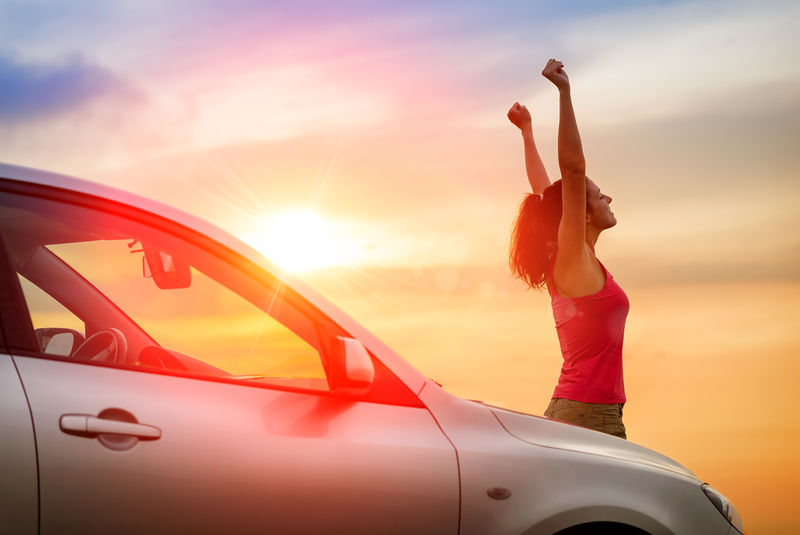 坐在车旁的女司机举起双臂-感受着朝着日落方向驾驶的感觉-妇女和车辆在美丽的阳光背景下