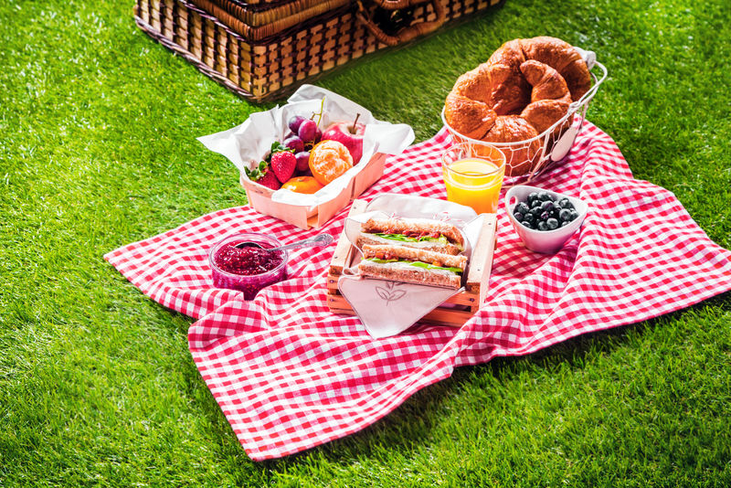 暑假的健康野餐-新鲜烤牛角面包、新鲜水果水果沙拉、三明治和一杯新鲜的橙汁放在红白格子布和篮子上