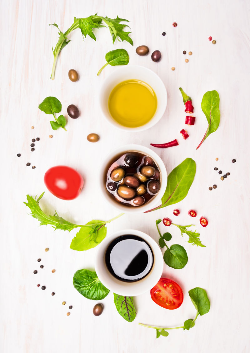 沙拉准备-配调味料、橄榄、野生香草叶、辣椒、油和番茄-白色木质背景-俯视图