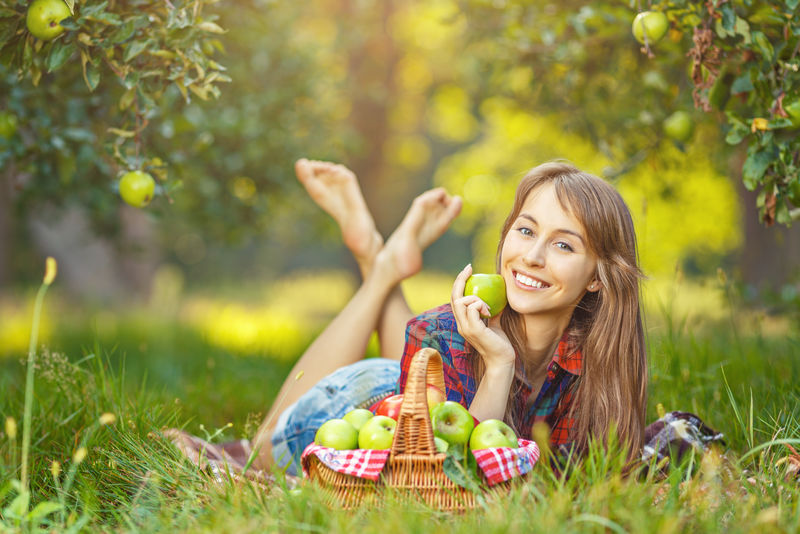 花园里有苹果的女人-一位年轻迷人的女士正躺在苹果园的草地上-微笑着-篮子里装满了成熟的红绿苹果-收获季节-乡村生活方式