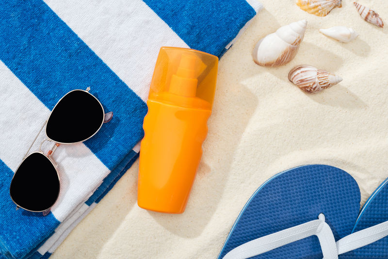 橙色的防晒霜，放在沙滩上，靠近条纹毛巾、蓝色人字拖、太阳镜和贝壳。