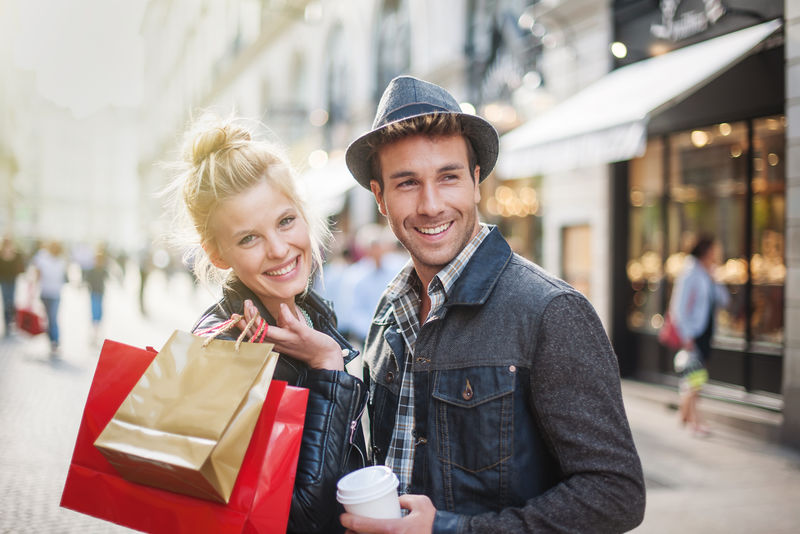 圣诞节期间-一对时髦的年轻夫妇在城里散步-年轻的女人穿着皮夹克-胳膊上挎着购物袋-男人手里拿着一杯咖啡