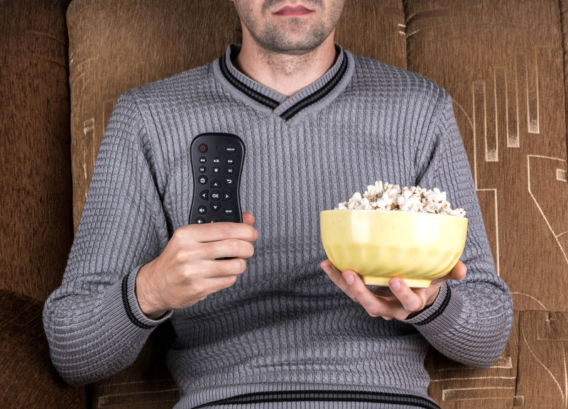 一个拿着遥控器和爆米花在沙发上看电视的男人