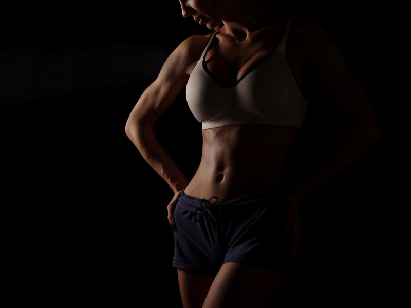 在演播室里穿着白色运动衫和蓝色短裤的年轻运动性感女性在黑色背景下展现了强壮的二头肌和平坦的腹部，完美的腹部肌肉，黑色背景