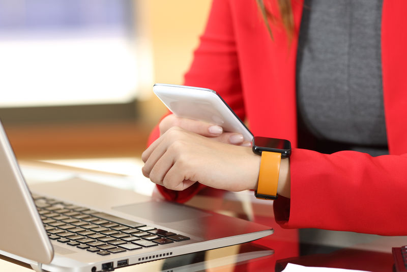 企业家双手同步智能手表和手机坐在办公室的桌面上-背景是她的红色西装
