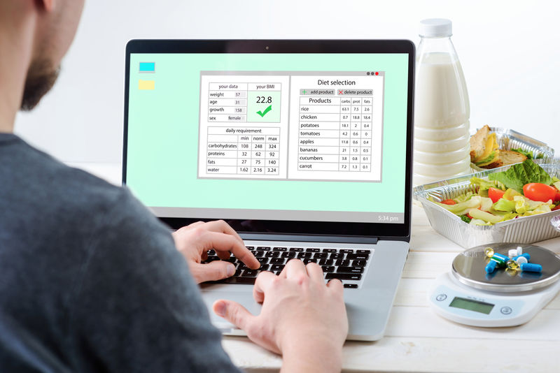 一个男人在笔记本电脑前-屏幕上有营养计划-特写-一瓶牛奶-沙拉-三明治-一个食物秤和药丸-白色背景-注册营养师