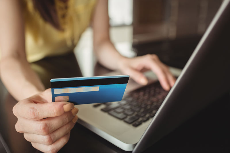 一位戴眼镜的年轻女商人拿着笔记本电脑坐在咖啡馆里-手里拿着信用卡在网上购物-网上购物的概念
