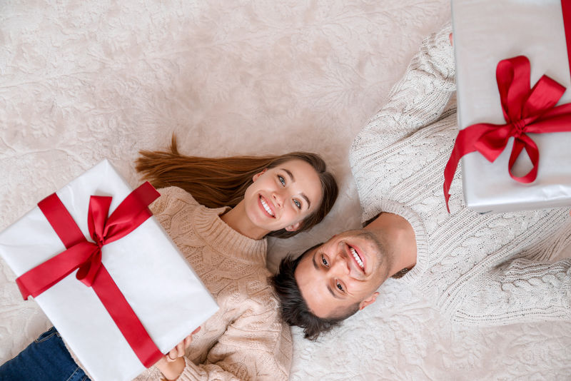 年轻幸福的夫妇躺在床上-西班牙裔男女拿着带丝带的礼物信封-周年庆典-卧室里有情人