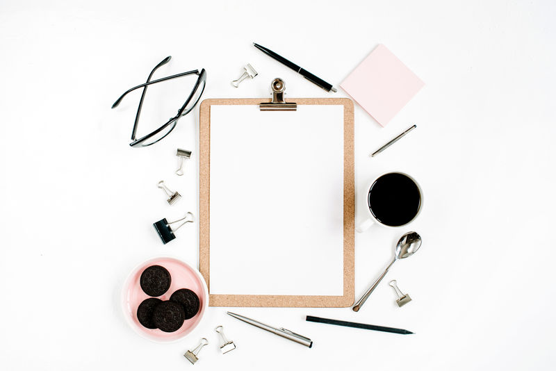 淡粉色的家庭办公室办公桌框架-带剪贴板、咖啡、饼干、眼镜和白色背景的办公用品-平放-俯视图-企业家办公室概念