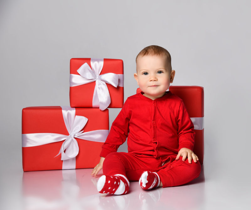 小男孩穿着圣诞老人的圣诞服装-戴着圣诞帽-开心地微笑着-带着礼物盒-准备在白色背景下庆祝新年派对