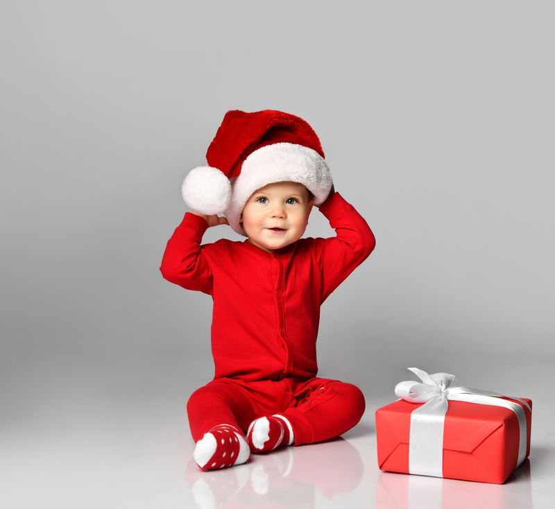 小男孩穿着圣诞老人的圣诞服装-戴着圣诞帽-开心地微笑着-带着礼物盒-准备在白色背景下庆祝新年派对
