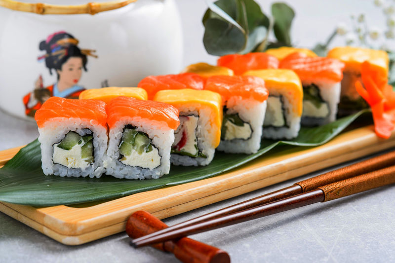 筷子夹寿司卷-配三文鱼、奶油奶酪和黄瓜-放在黑色石板上-Uramaki、Nori Maki或Futomaki寿司配鳟鱼片、酱油和芥末