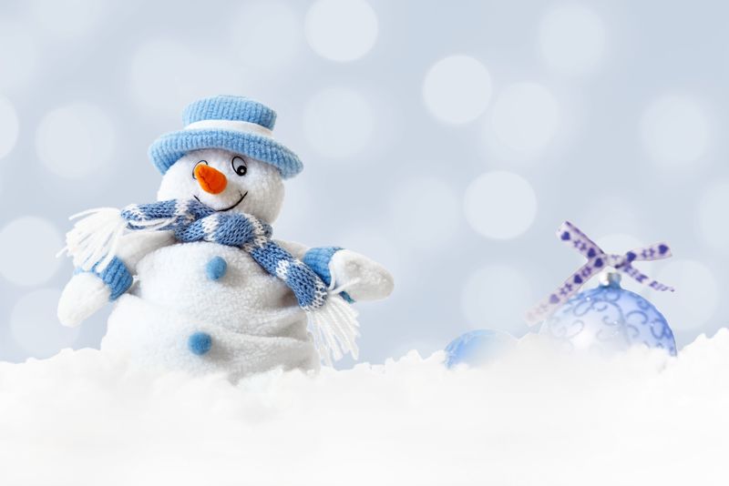 圣诞快乐的雪人戴着蓝色的帽子和围巾-抽象的灯光背景上挂着圣诞球-冬天的风景上飘落着白色的雪花-圣诞快乐和新年快乐的贺卡