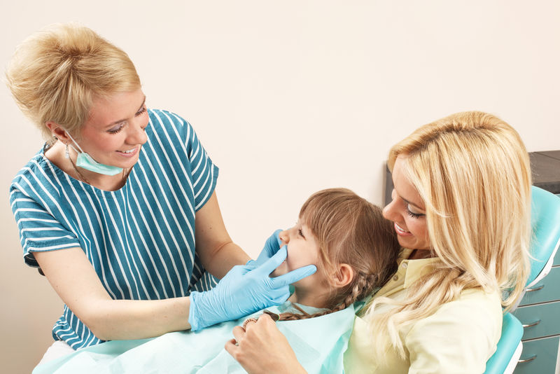 小女孩坐在妈妈的膝上，牙医检查她的牙齿