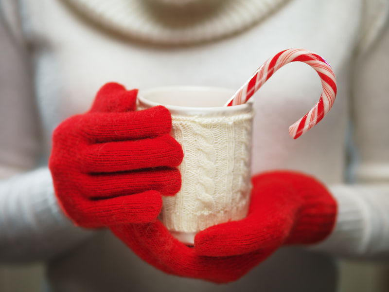 一名妇女双手戴着毛红色手套，手里拿着舒适的杯子，里面有热可可、茶或咖啡和一根甘蔗。冬季和圣诞节的概念。
