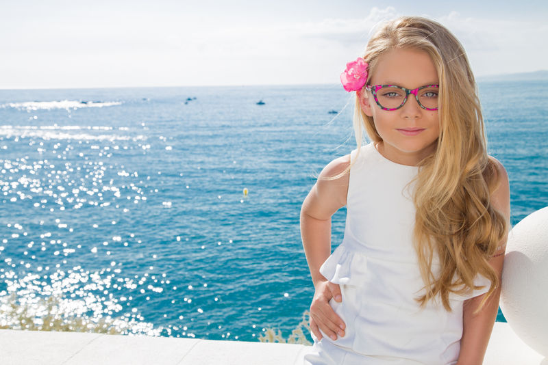 在西班牙、希腊、圣托里尼，年轻漂亮的女孩戴着粉色眼镜、穿着别致的连衣裙，梳着金色卷发，站在水池边，伴着栏杆、岩石和大海