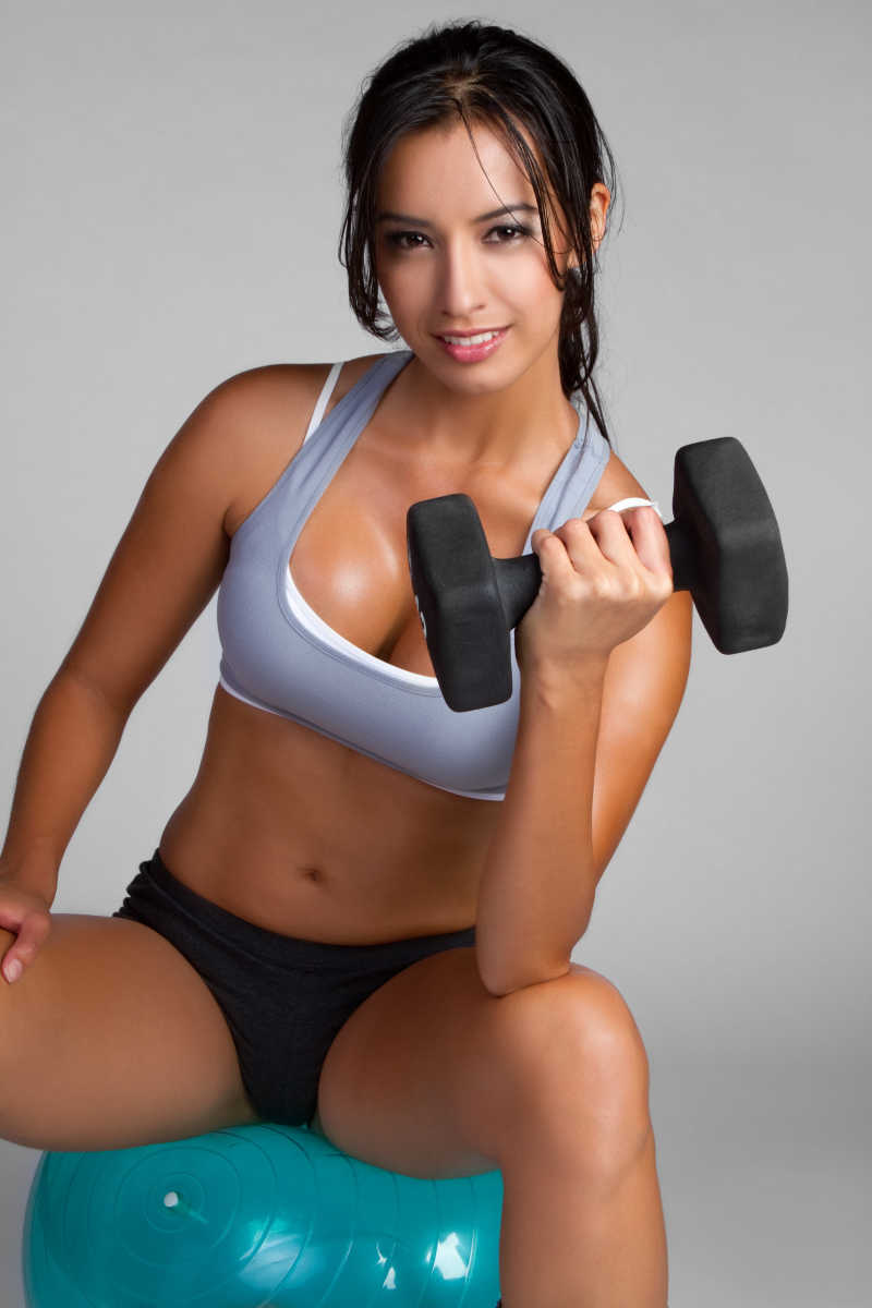 灰色背景上坐在瑜伽球上用哑铃锻炼的健身女人