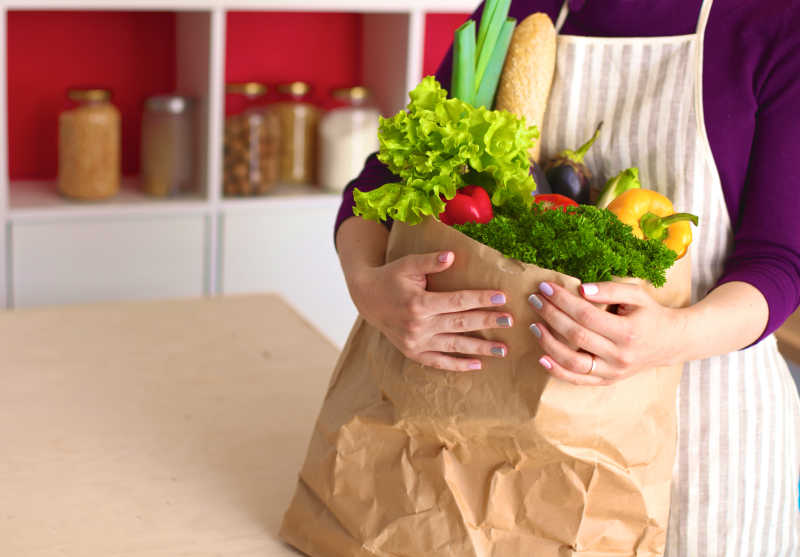 女人手里拿着一个满是和蔬菜的纸质购物袋