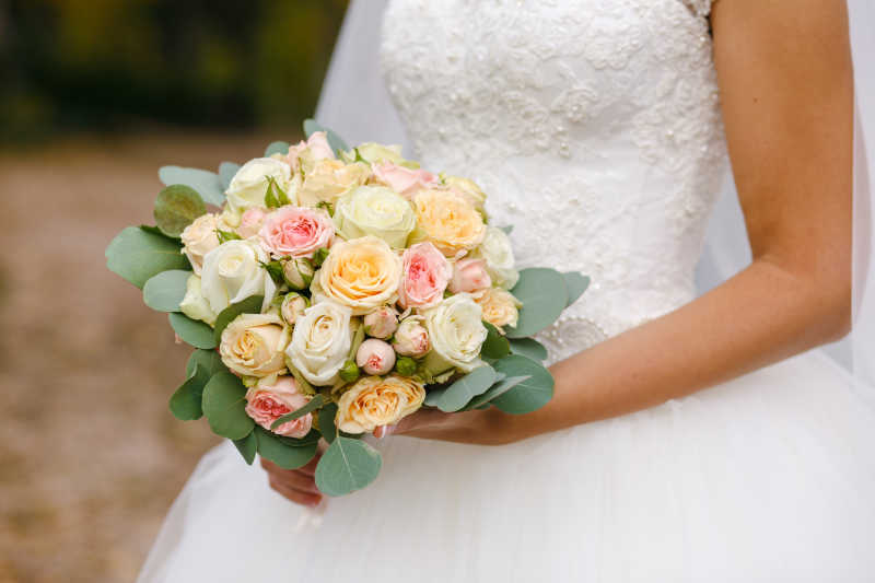 浅黄色户外背景下的穿着白色婚纱的新娘手中的美丽婚礼花束