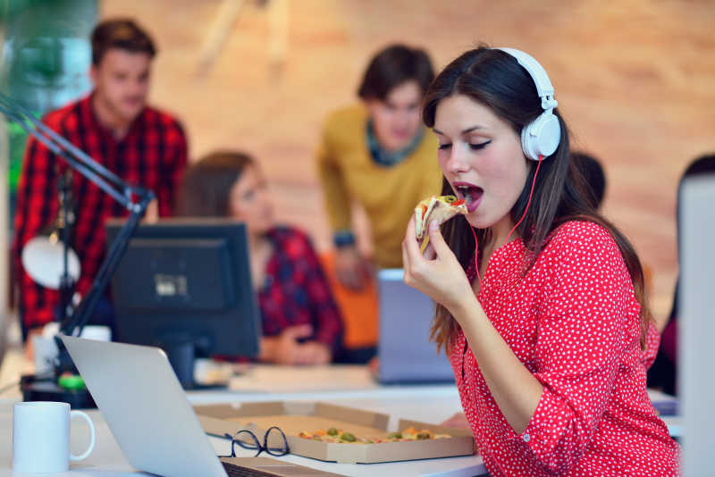 现代化办公室背景下带着耳机吃披萨的美女