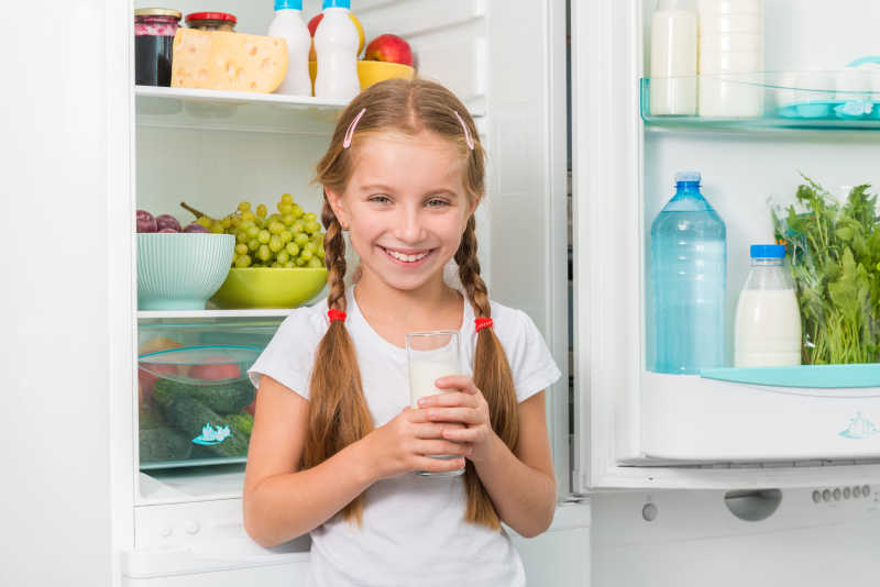冰箱旁边的小女孩拿着一杯牛奶