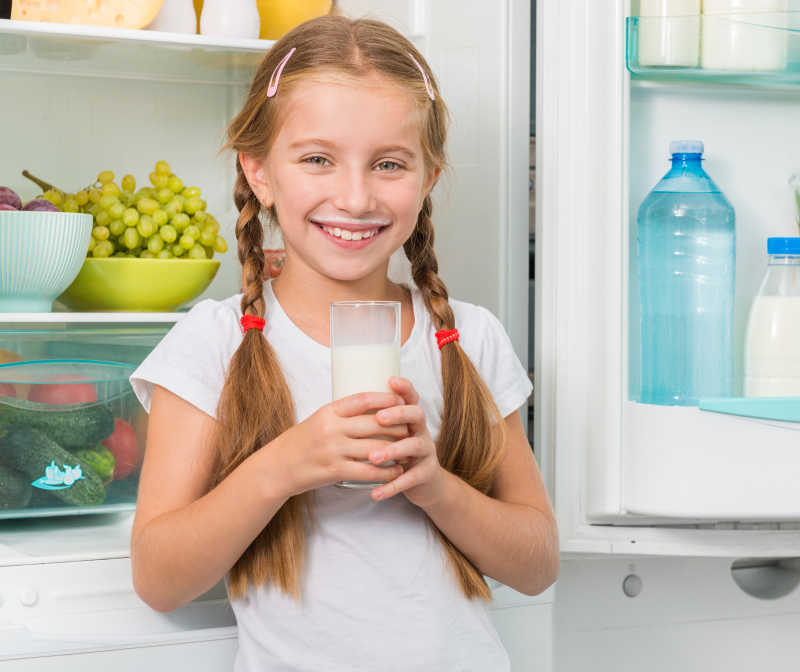 冰箱旁边拿着一杯牛奶的小女孩