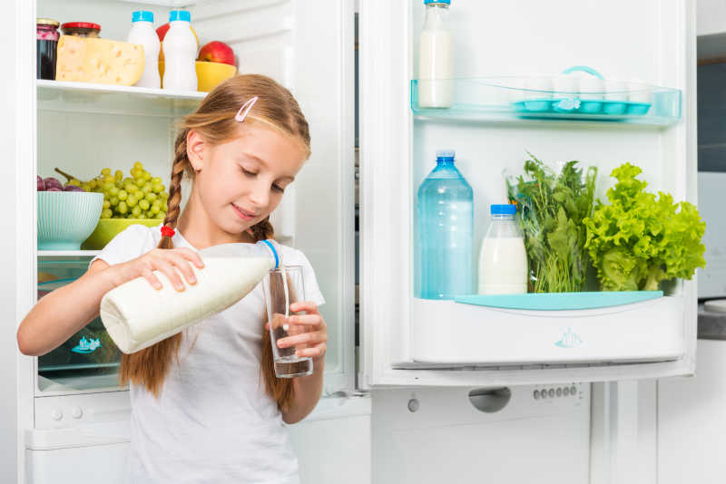 冰箱旁边的小女孩把牛奶倒在玻璃杯里
