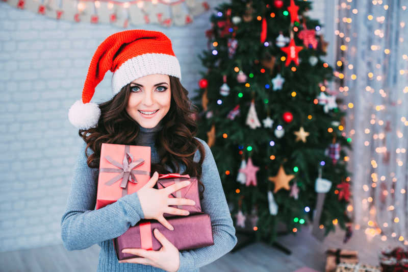圣诞树背景前抱着圣诞礼盒微笑的美女
