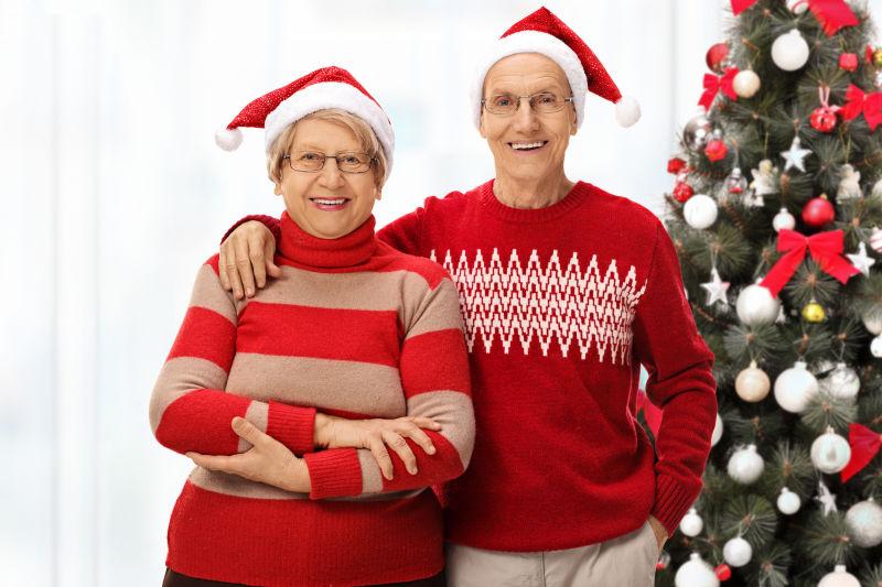 站在圣诞树前的带着圣诞帽子的快乐老夫妇