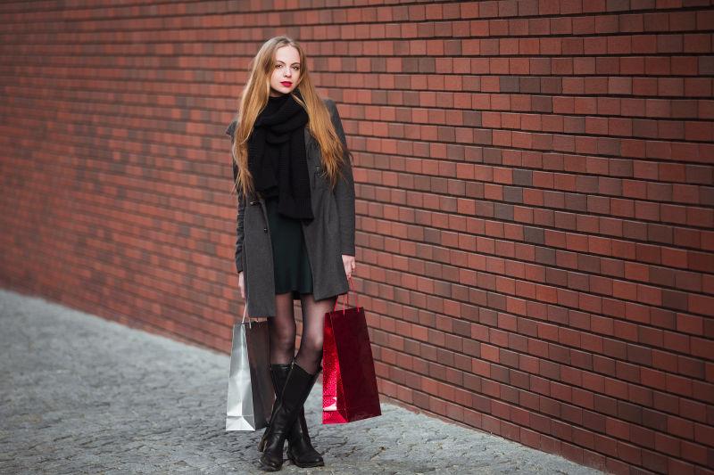暗红色砖墙背景前提着购物袋的黑色风衣美女