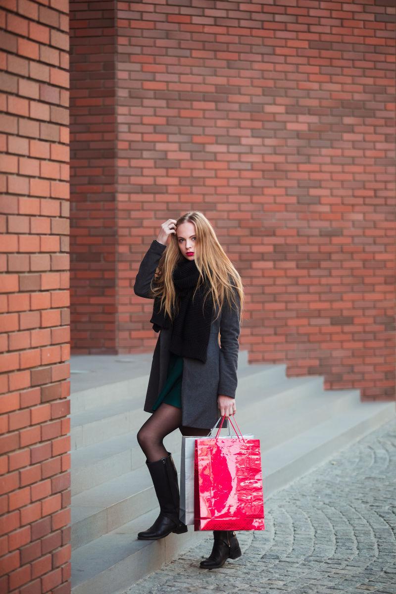 砖红色建筑前穿着黑色风衣拎着购物袋上楼的美女